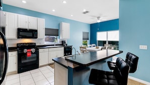 A modern kitchen with white cabinets, a black microwave, and a large island with black bar stools. - Aster Cove (Kissimmee)