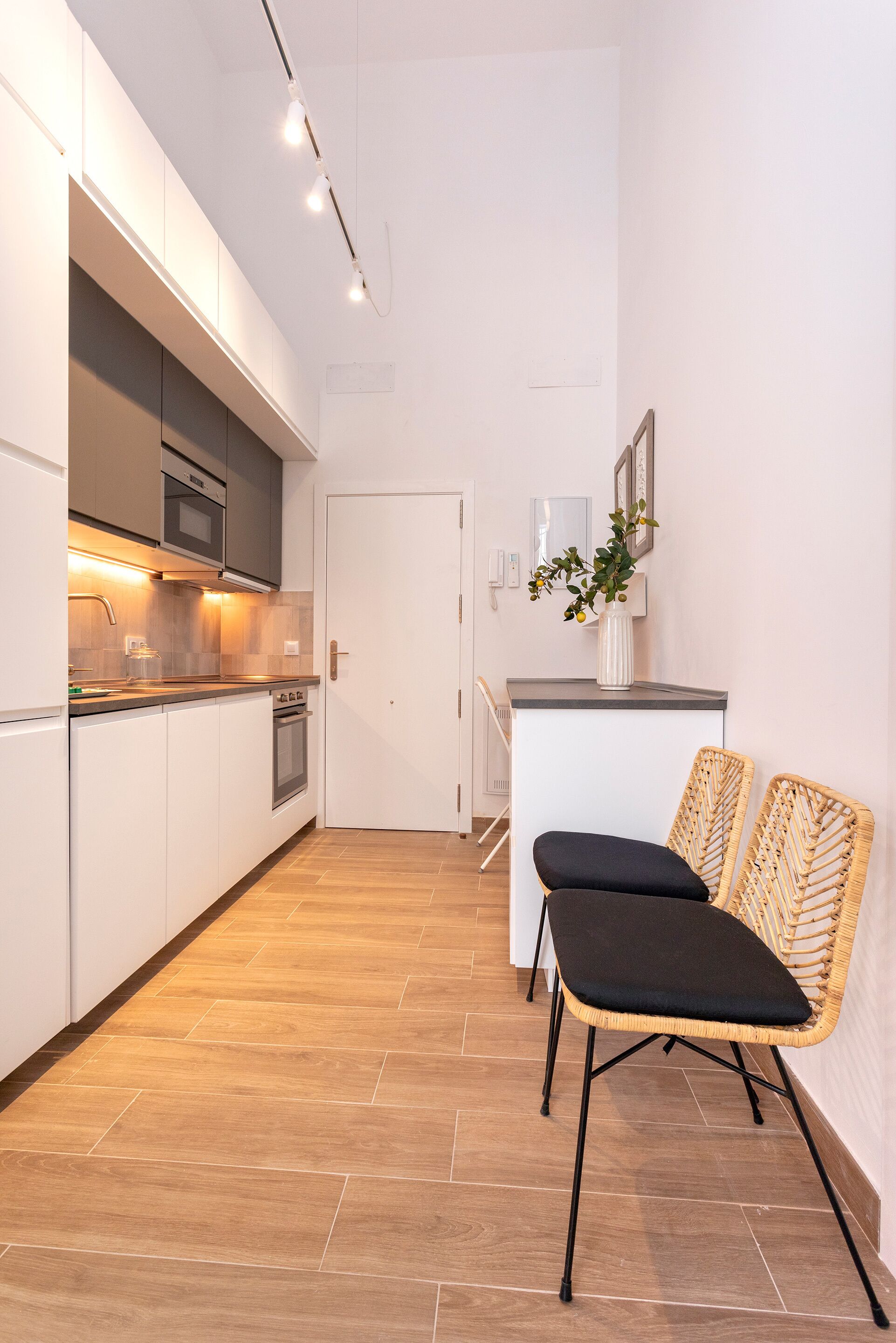 Una cocina moderna con armarios blancos, horno empotrado y microondas. Hay una zona de comedor con dos sillas de mimbre y una mesita.