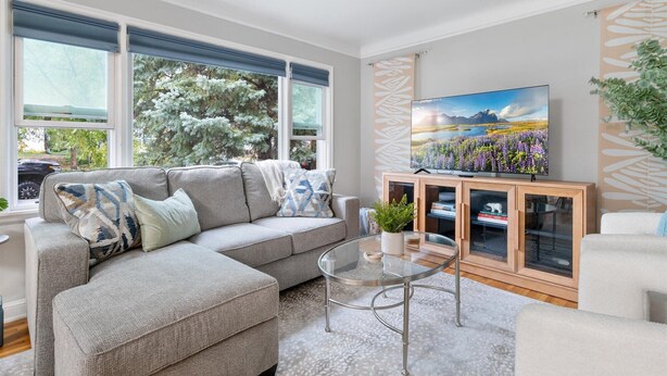 A living room with a sectional sofa, a glass coffee table, a TV on a wooden cabinet, and a view of trees through large windows. - Theater - Firepit & BBQ - Life Size Games (Minneapolis)