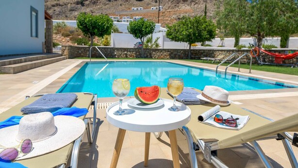Un espace aménagé au bord de la piscine, avec des chaises longues, une table sur laquelle on trouve une pastèque et des boissons, et une vue sur un paysage montagneux.
