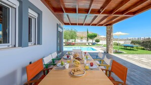 Un espace de restauration en plein air avec vue sur la piscine, comprenant une table en bois, des chaises et une variété d’aliments pour le petit-déjeuner.