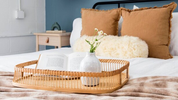 Een netjes opgemaakt bed met een witte deken, een bruin kussen en een zacht, wit kussen. Een rieten dienblad met een witte vaas en een boek staat op het bed.
