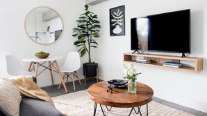 Een moderne woonkamer met een ronde houten salontafel, een bank, een eettafel met stoelen, een grote potplant, een flatscreen televisie op een houten dressoir, en een spiegel aan de muur.