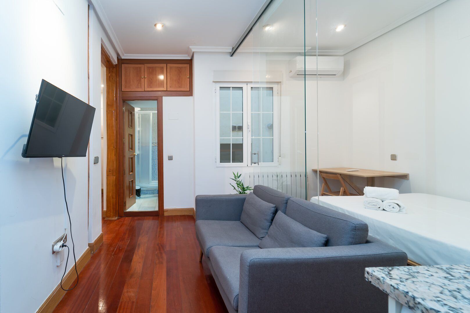 Una habitación moderna con un sofá gris, una mesita, un televisor montado en la pared y una ventana con persianas.