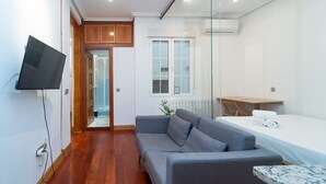 Ett modernt rum med en grå soffa, ett litet bord, en tv som är monterad på väggen och ett fönster med persienner.