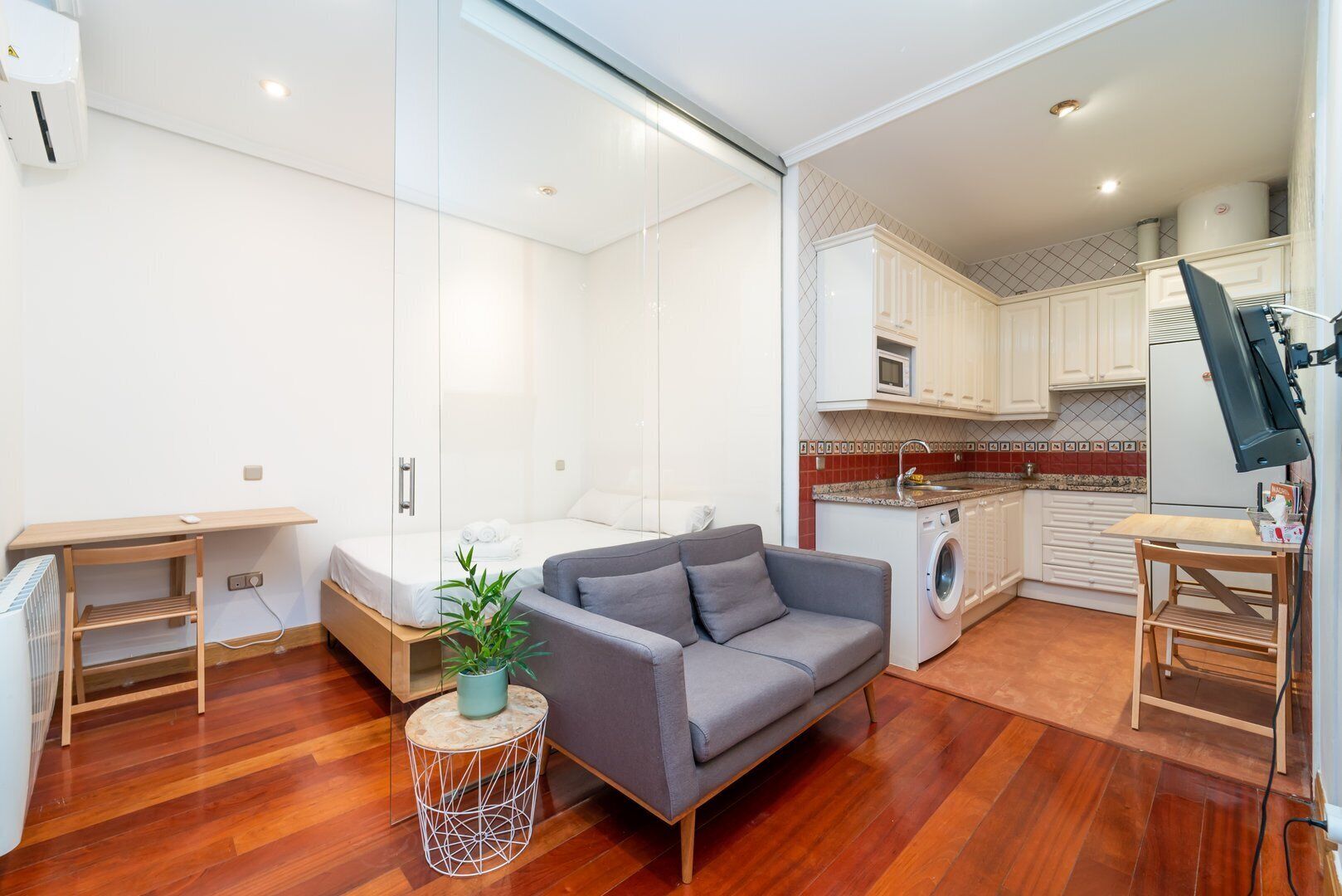 A modern apartment with a grey sofa, a small round table with a plant, a washing machine, and a kitchen with white cabinets.