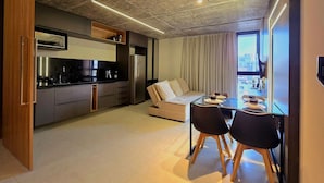 A modern kitchen with a built-in oven and microwave, a dining area with a glass table and chairs, and a sofa with a view of the city through large windows. - Urban by Hosppedar (João Pessoa)