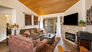 Una sala de estar acogedora con techo de madera, chimenea, televisor de pantalla plana y un gran ventanal con vistas al exterior.