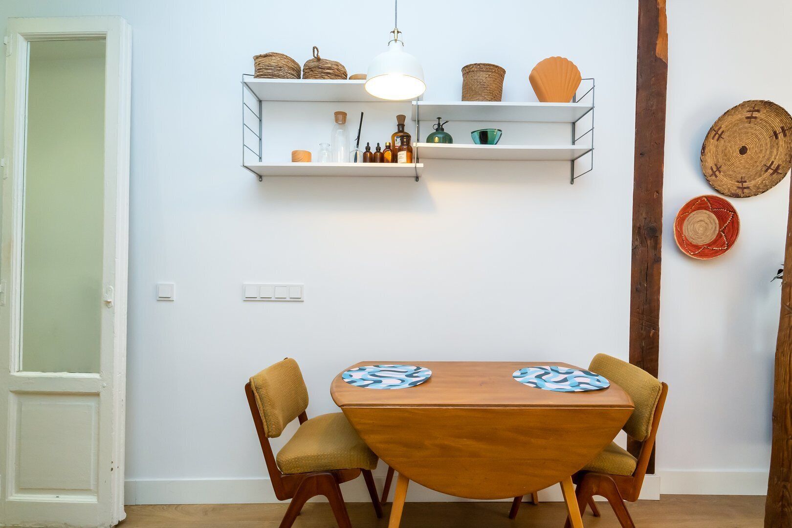 Un espace repas comprenant une table et des chaises en bois, un luminaire suspendu et des étagères ornées d’objets décoratifs.
