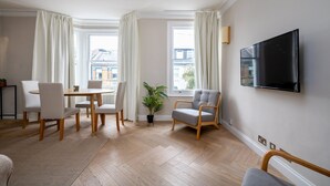 A modern living room with a dining area, large windows, a flat-screen TV, and a potted plant. - GuestReady - Charming residence in Fulham (London)