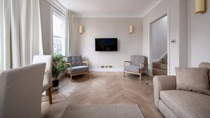 A modern living room with a large TV, two grey armchairs, a white sofa, and a potted plant. - GuestReady - Charming residence in Fulham (London)