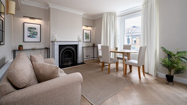 A living room with a fireplace, a dining area with chairs, and a large window with curtains. - GuestReady - Charming residence in Fulham (London)