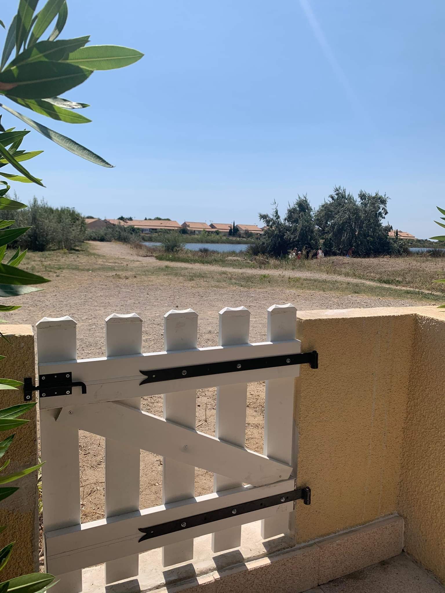 A white metal gate with a black latch, set against a beige wall, overlooking a grassy area with a body of water and buildings in the distance.