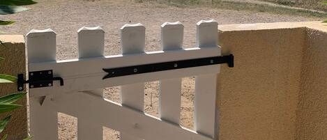 A white metal gate with a black latch, set against a beige wall, overlooking a grassy area with a body of water and buildings in the distance.