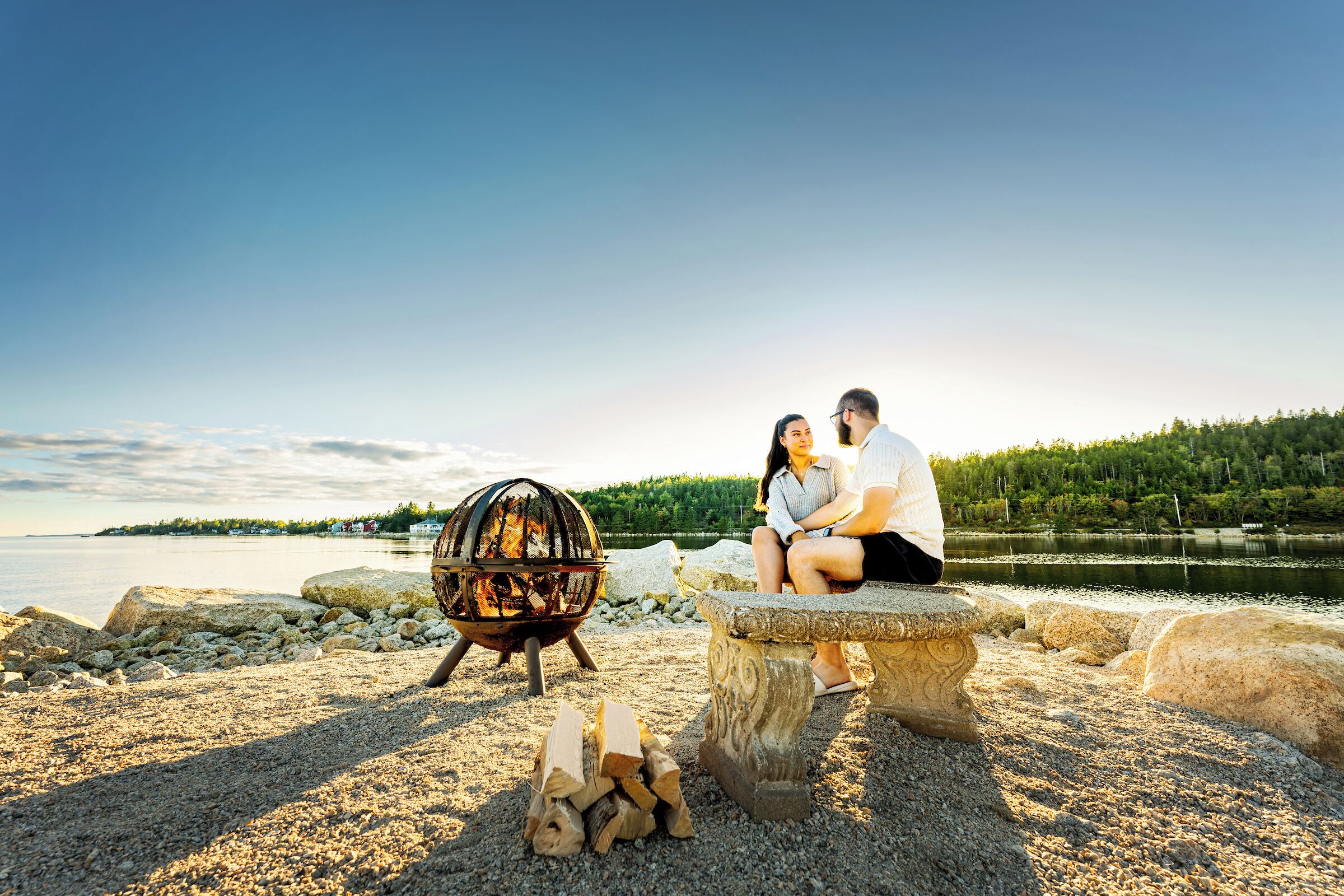 Una pareja sentada en un banco de piedra junto a un lago, con un brasero y leña cerca.