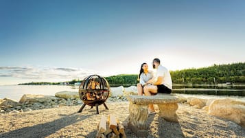 Una pareja sentada en un banco de piedra junto a un lago, con un brasero y leña cerca.