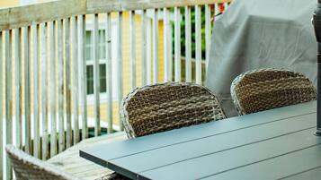 A patio with a table and wicker chairs, a covered grill, and a view of a house and trees.