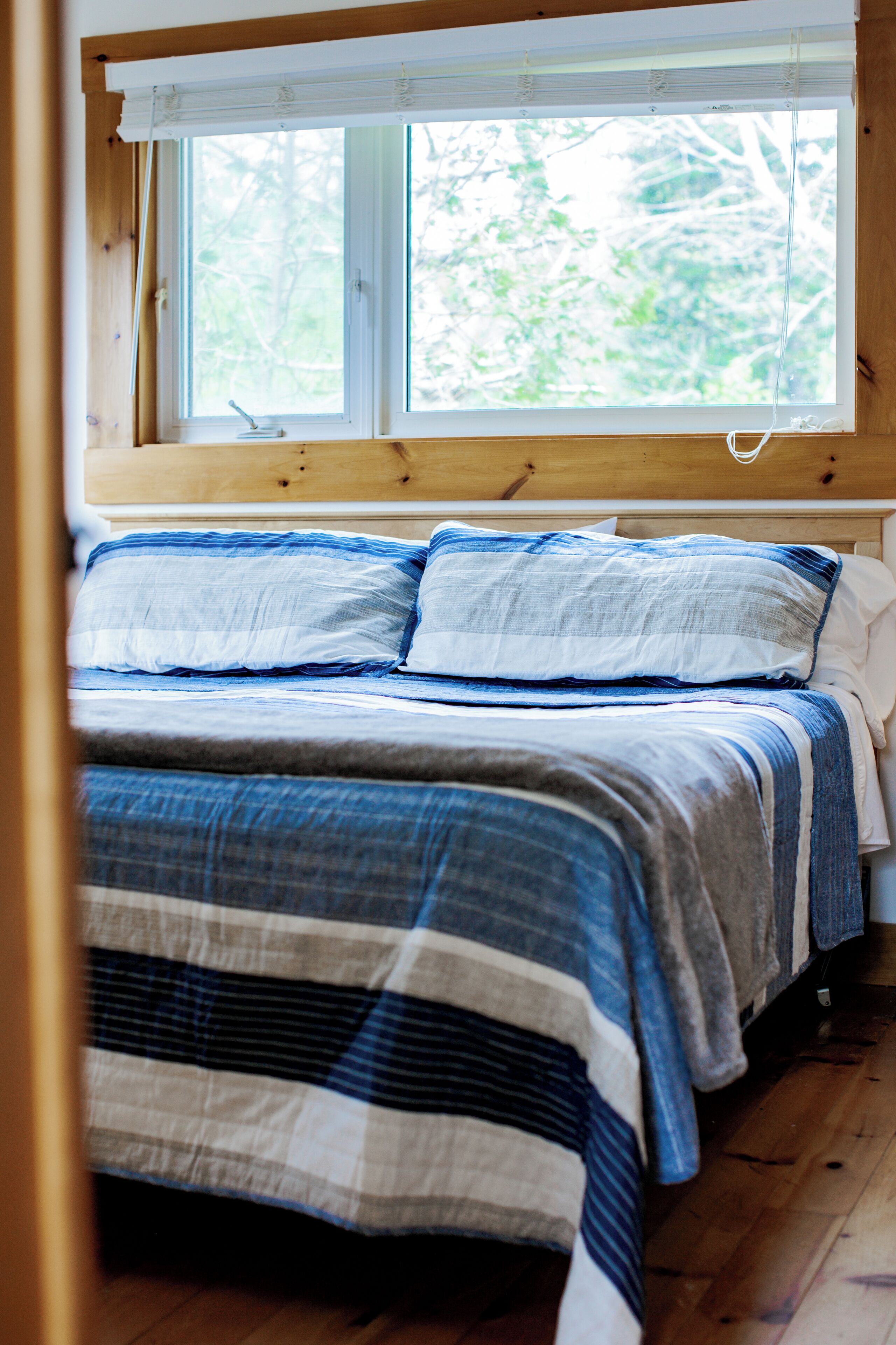 A bed with a striped blanket and pillows, a wooden headboard, and a window with a view of trees.