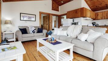 Un salon chaleureux avec un plancher en bois, une table basse blanche, un canapé gris et un coin cuisine doté de meubles en bois et d’un réfrigérateur blanc.