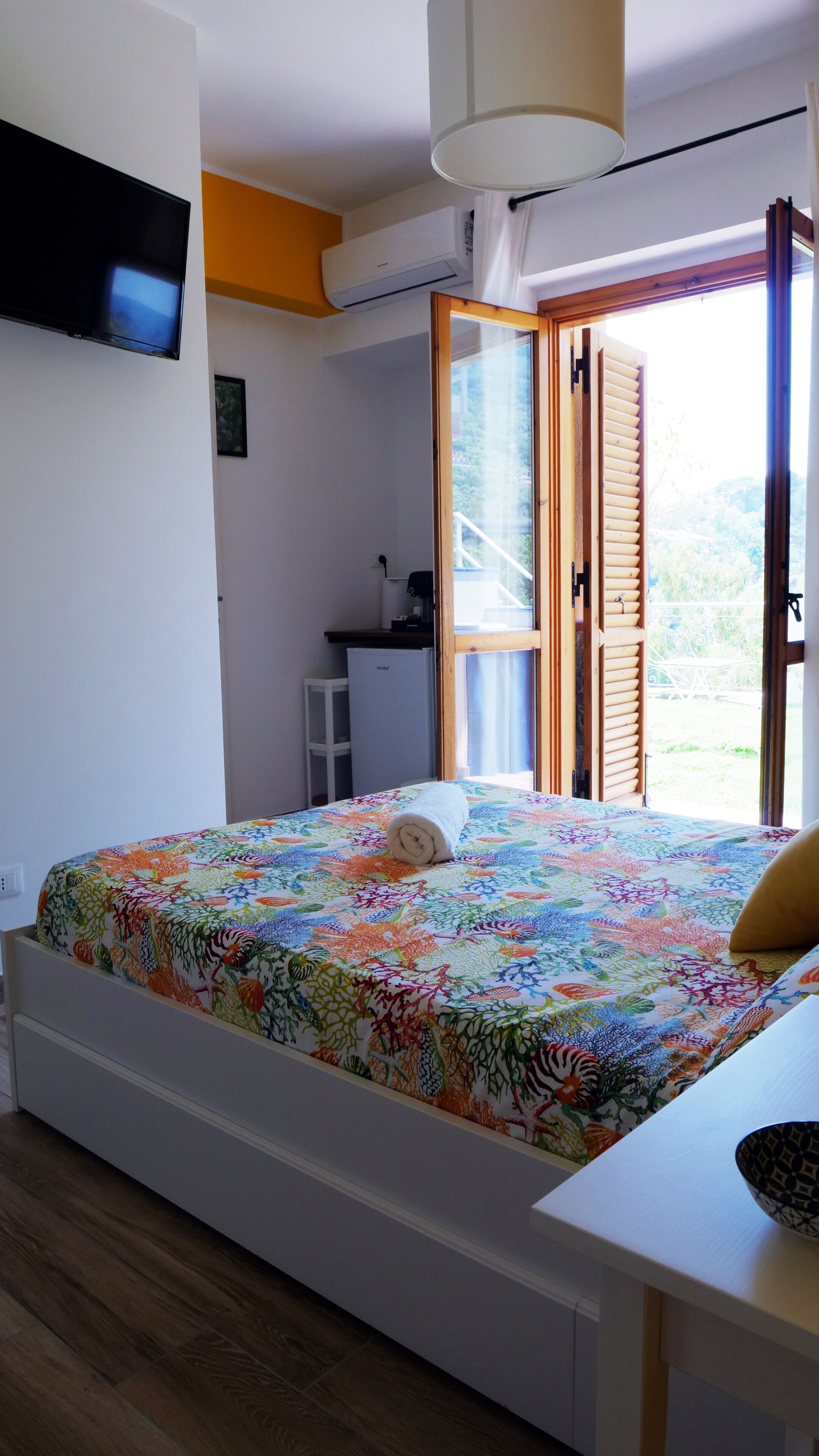 A bedroom with a bed, a desk, a TV, and a window with a view of greenery.