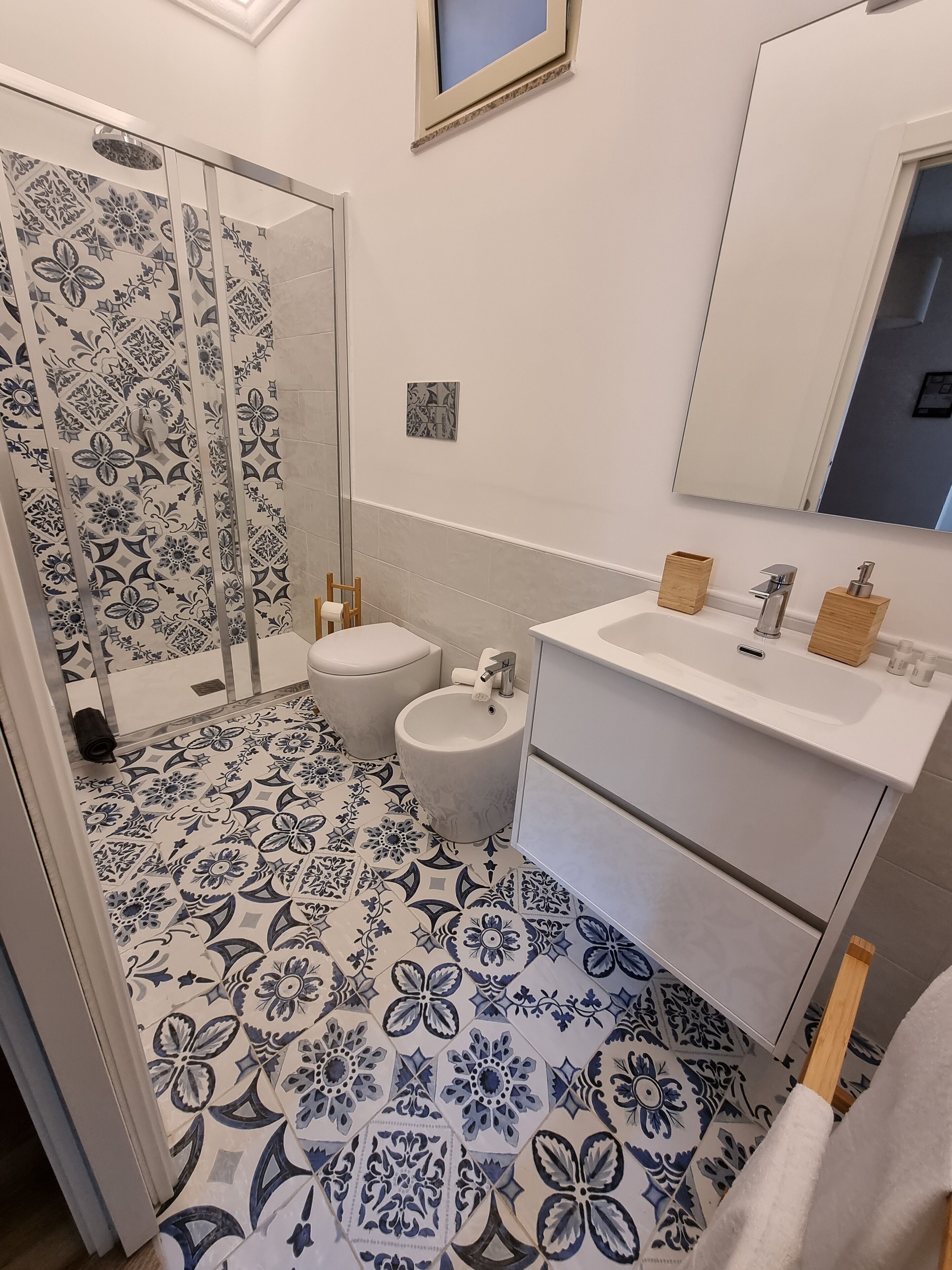 A bathroom with patterned floor tiles, a glass shower enclosure, a toilet, a bidet, and a sink with wooden soap holders.