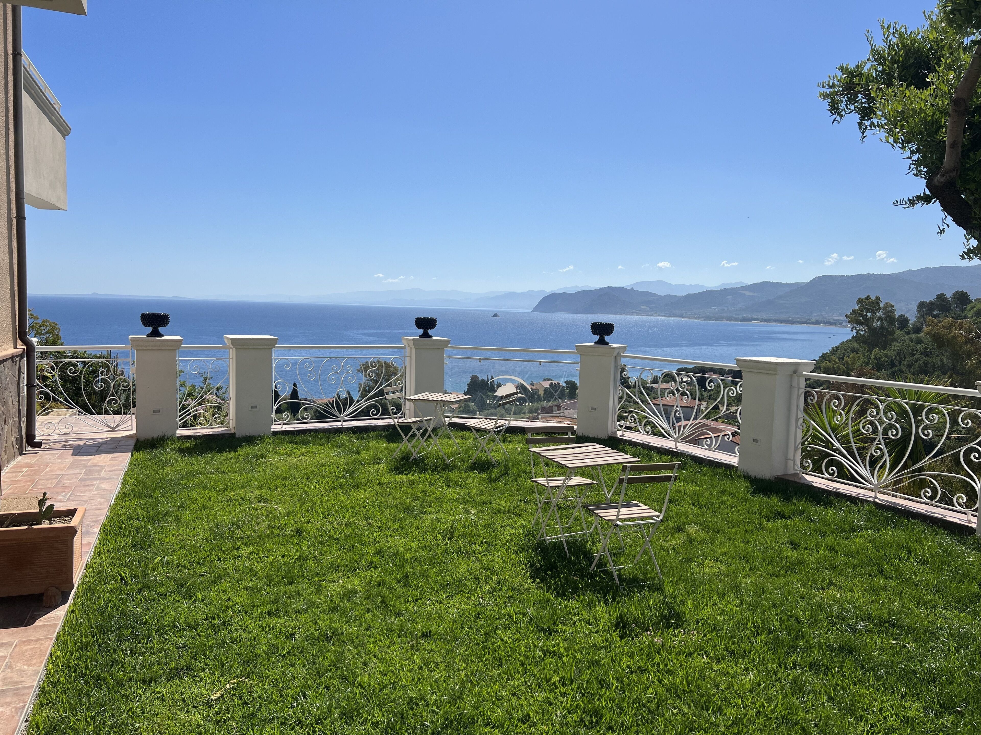 Un balcón con vista al mar, un área verde y una barandilla blanca.