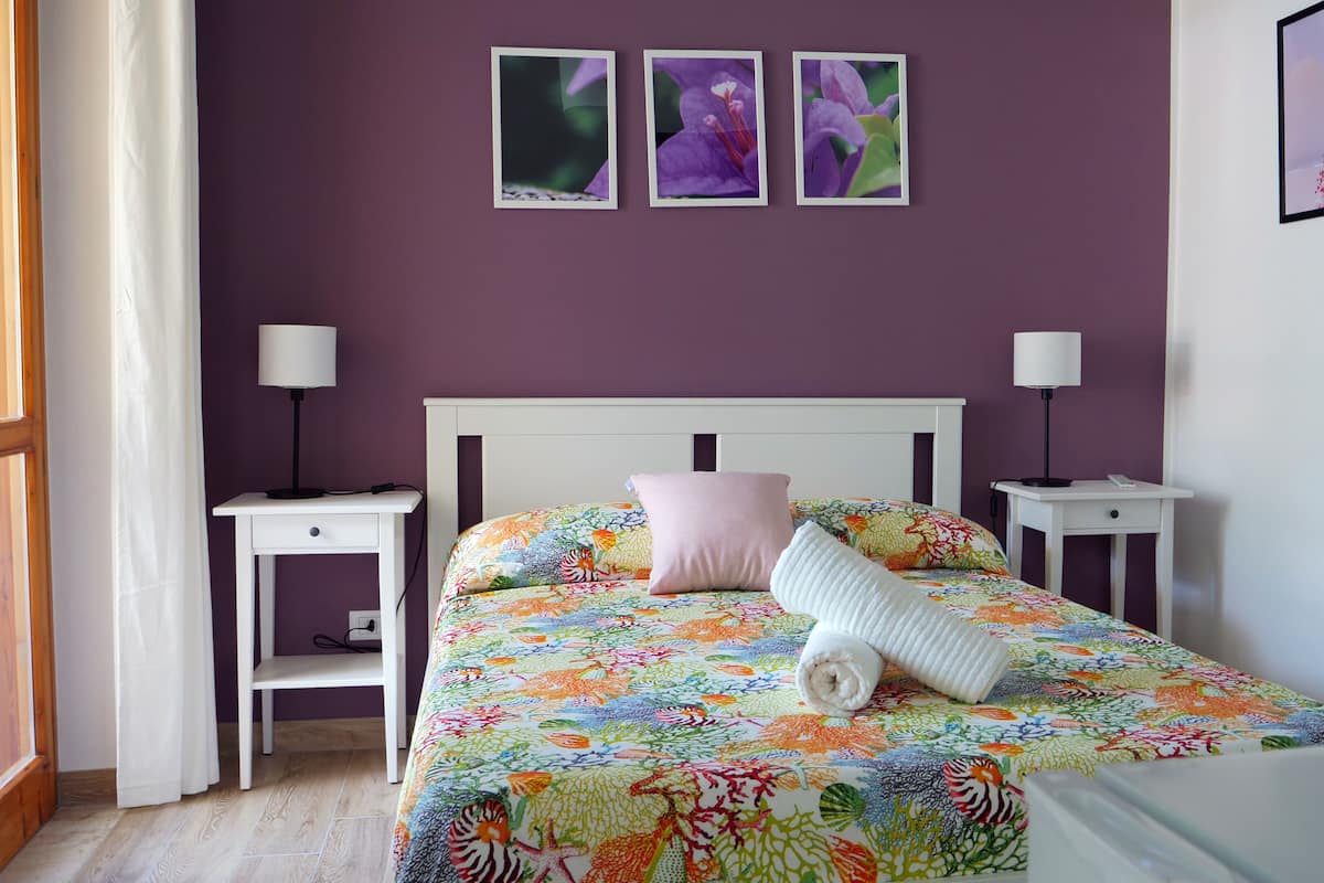 A bedroom with a bed, two bedside tables, a nightstand, a window with curtains, and three framed pictures on the wall.