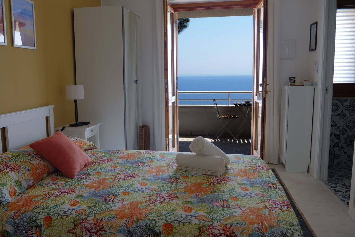 A bedroom with a bed, a nightstand, a lamp, a window with a view of the sea, and a balcony with a chair and table.