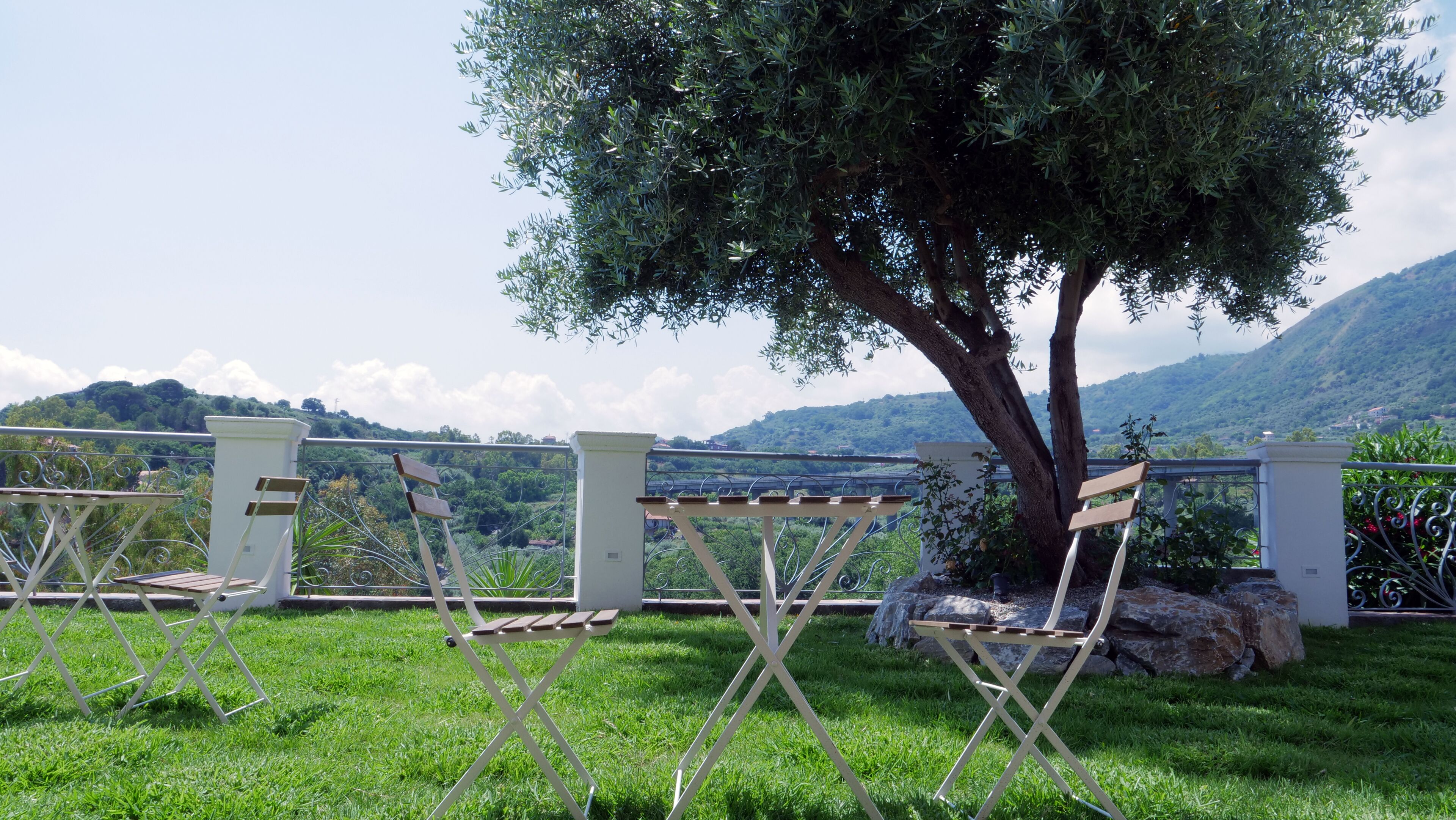 Un espace extérieur aménagé avec des chaises pliantes et une table, entouré d’une clôture blanche, et offrant une vue sur une colline et un arbre.