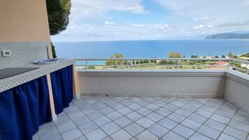 A balcony with a tiled floor, a blue curtain, and a view of the sea and distant hills.