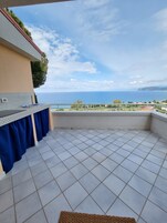 A balcony with a tiled floor, a blue curtain, and a view of the sea and distant hills.