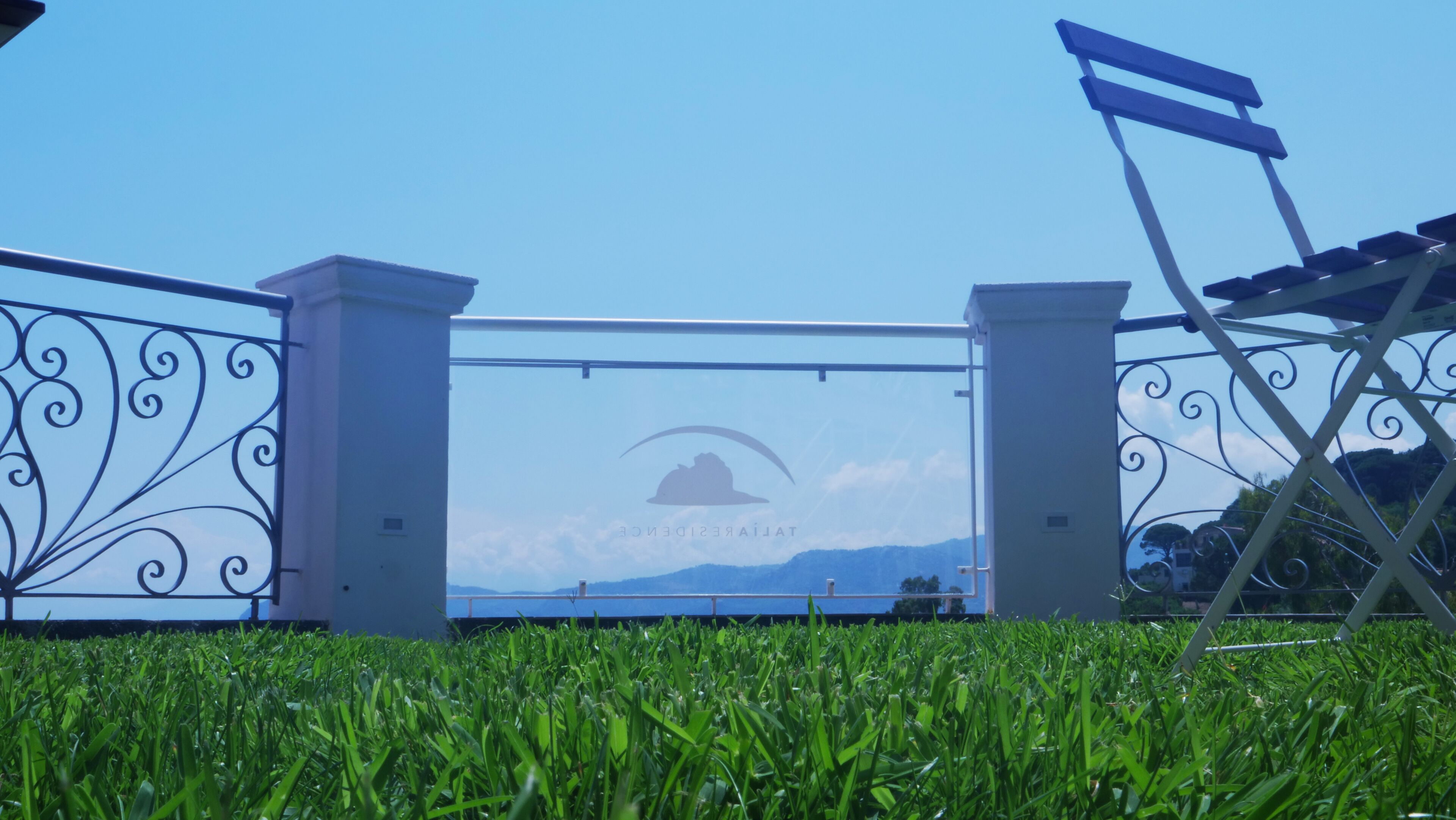 Een metalen poort met een logo, een grasveld voor en een heldere lucht.