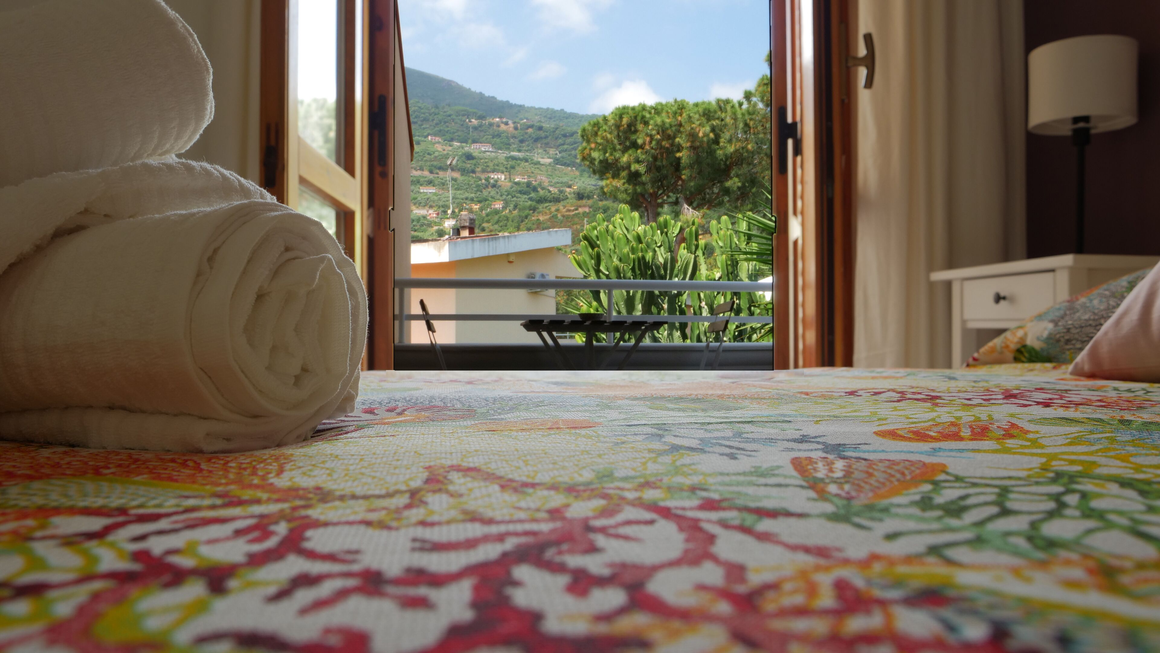 A bed with a colorful bedspread and a rolled towel on it. There is a window with a view of greenery and buildings.