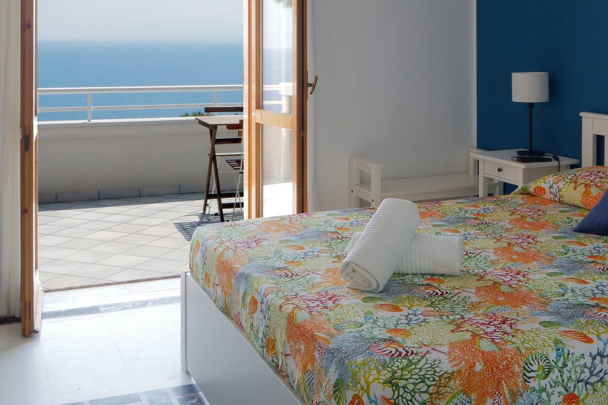 A bedroom with a bed, a nightstand, a lamp, a picture on the wall, and a view of the sea through an open door.