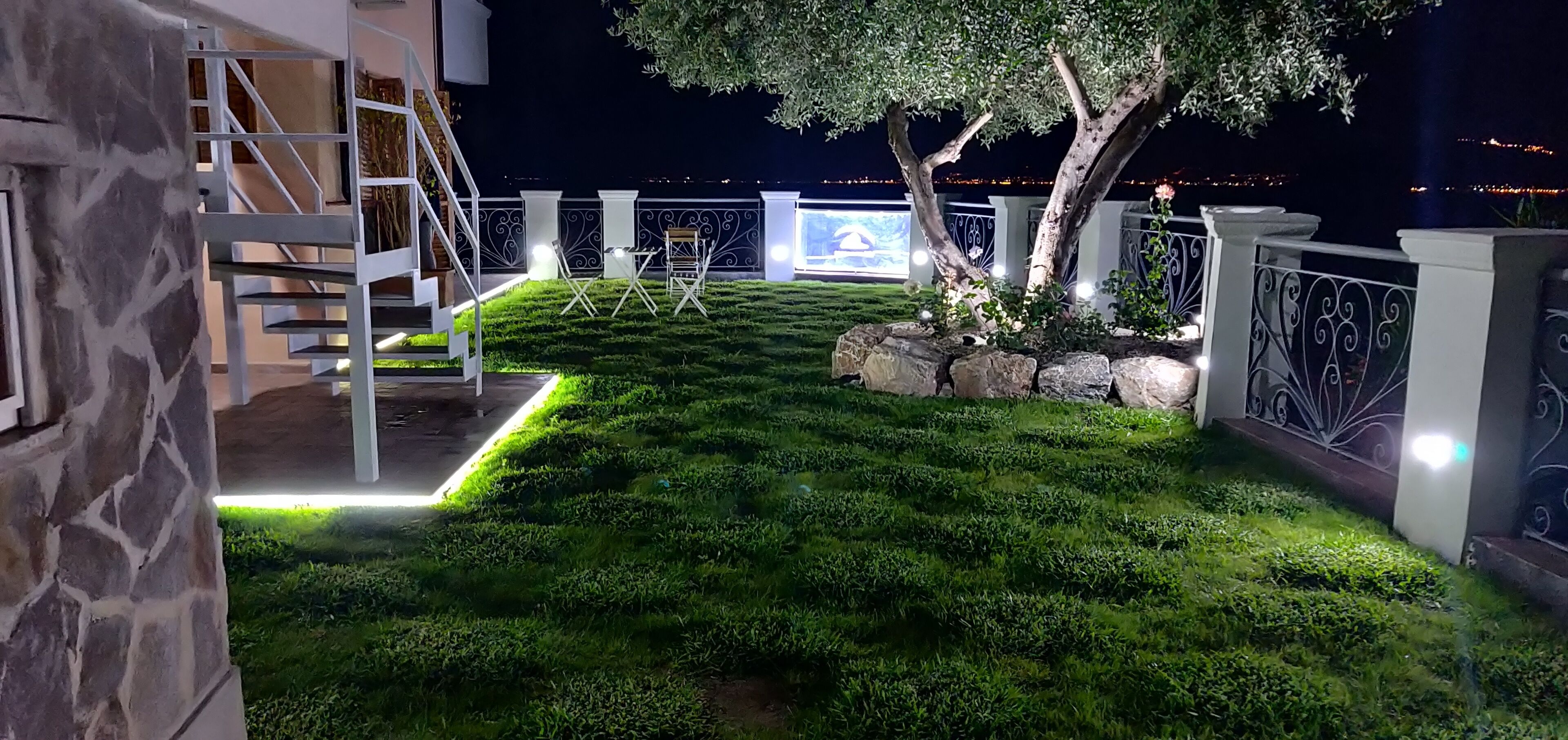 Un espace extérieur bien éclairé, avec un mur en pierre, un escalier en métal et une pelouse verdoyante agrémentée d’un arbre.