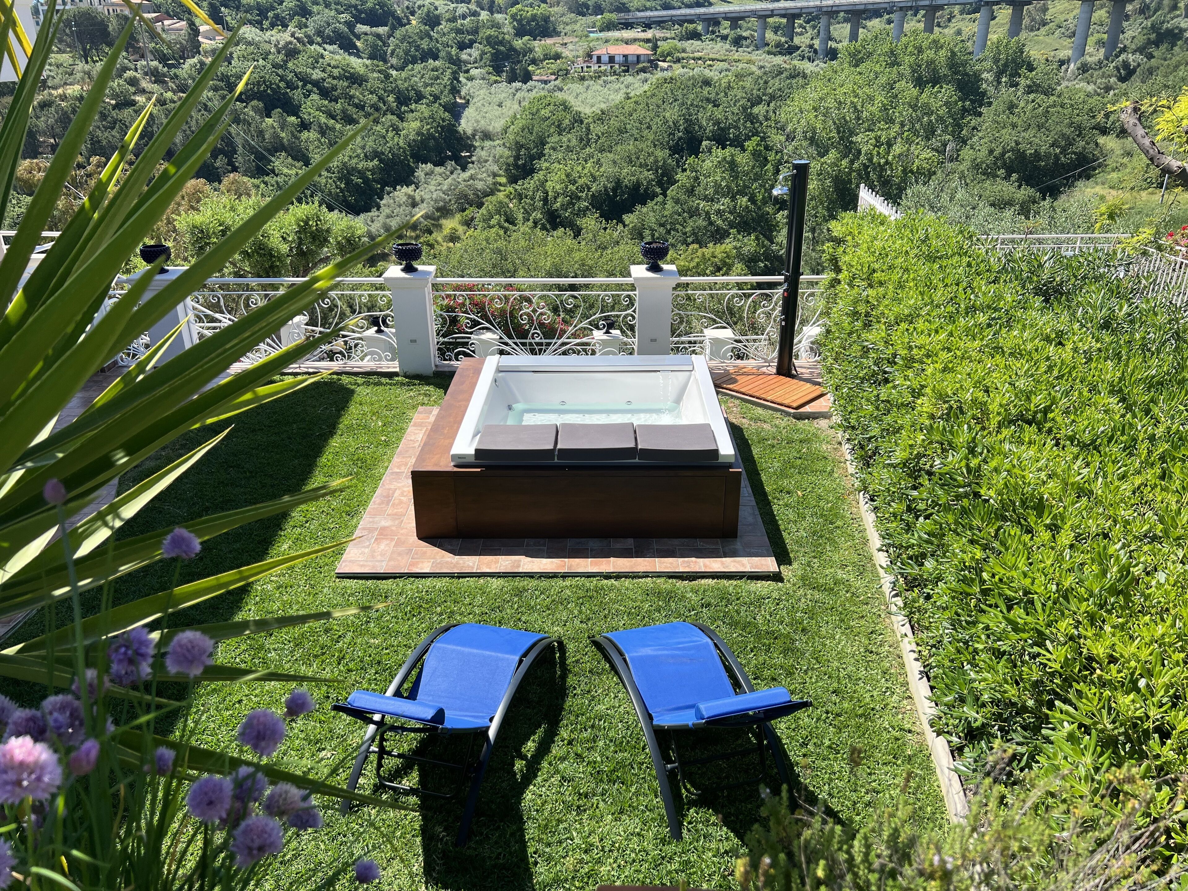 A hot tub with two blue lounge chairs in a garden with a view of a bridge and hills.