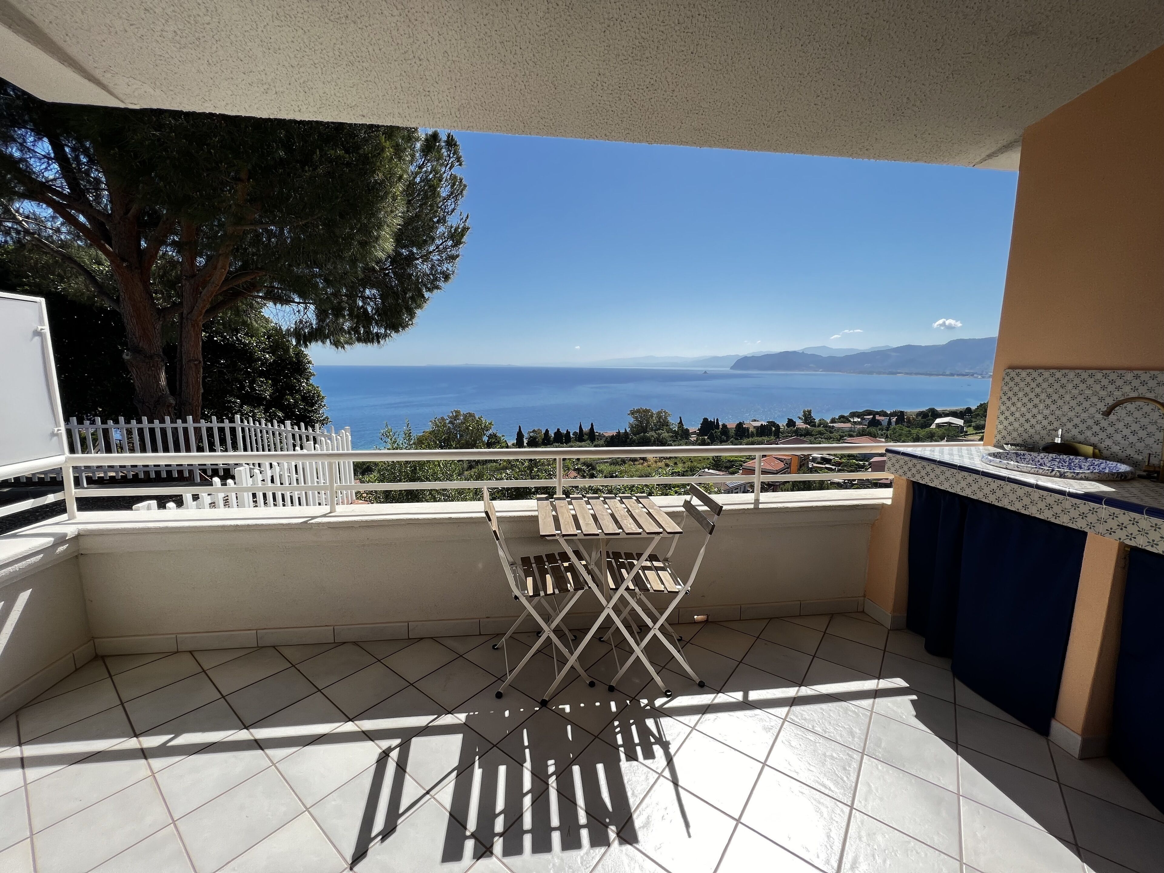A balcony with a view of the sea, a white fence, and a tiled floor.