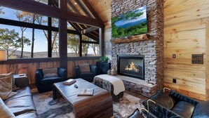 A cozy cabin interior with a stone fireplace, wooden walls, and a large window overlooking a forested area.