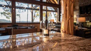 A marble table with a bottle of Crema wine and three glasses. The setting includes wooden beams, a stone wall, and a view of trees through large windows.