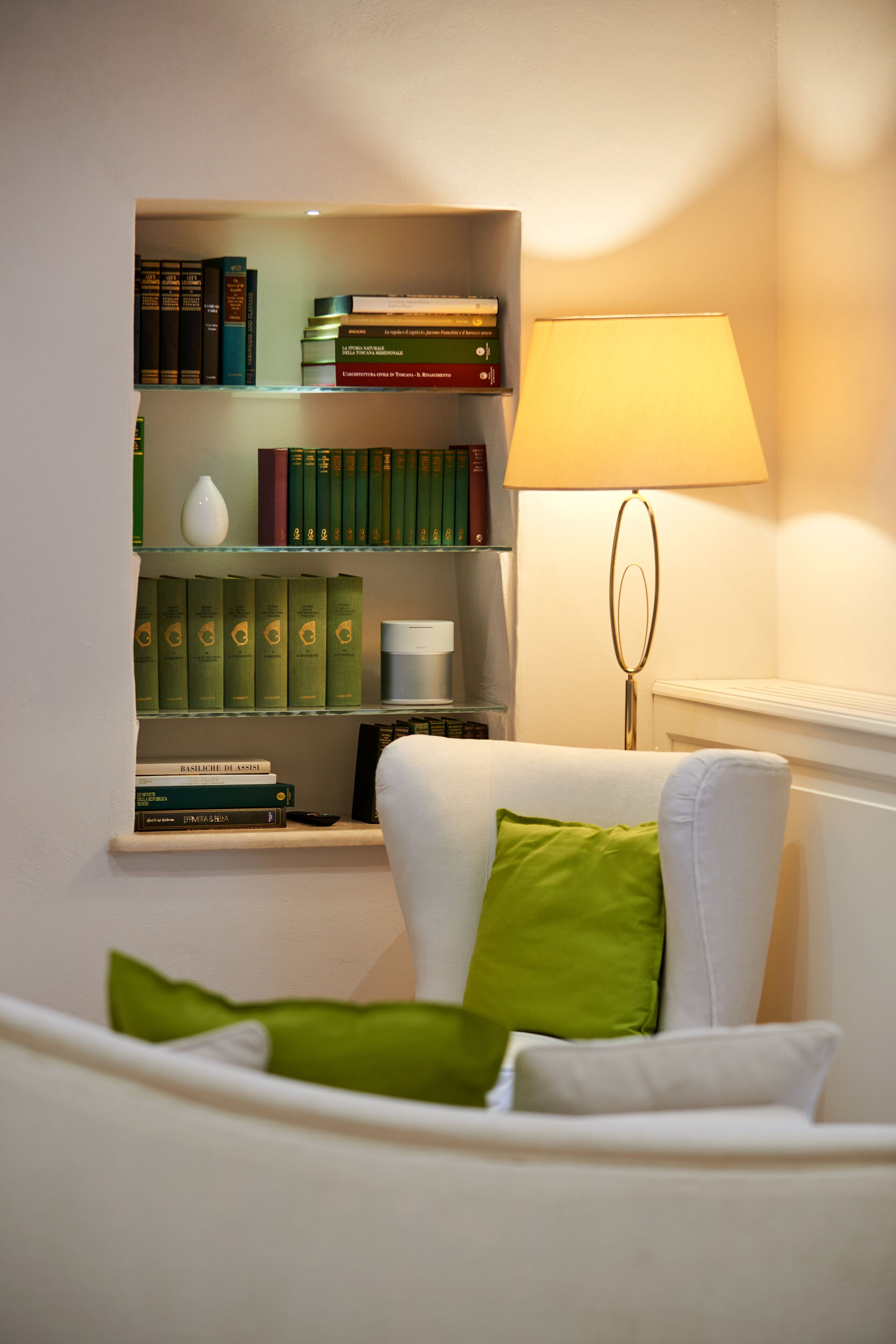 A living room with a white sofa, green cushions, a bookshelf with books and decorative items, and a floor lamp.