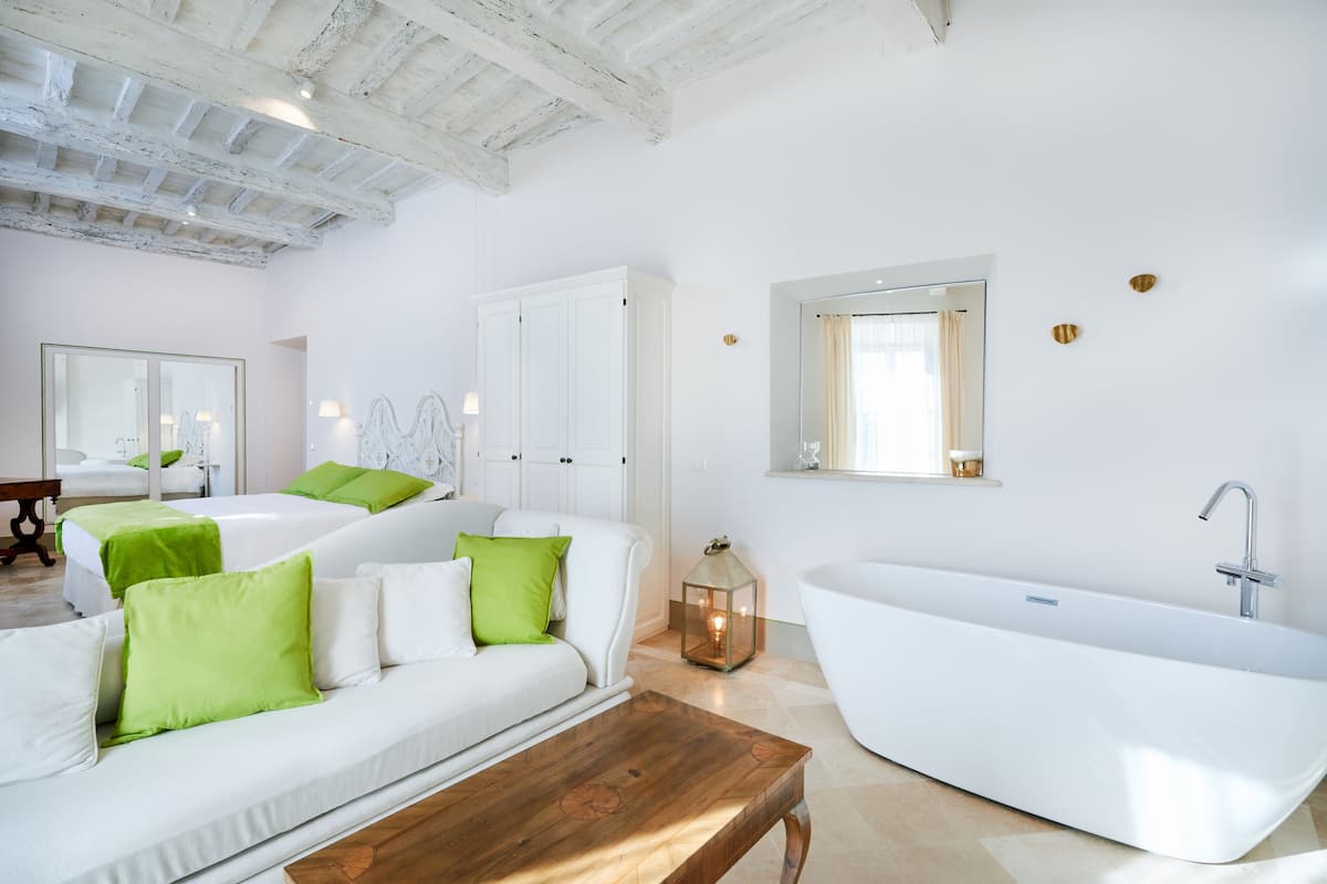 Een moderne hotelkamer met een wit kleurenpalet, een groot ligbad, een houten bijzettafeltje en een groen sierkussen.