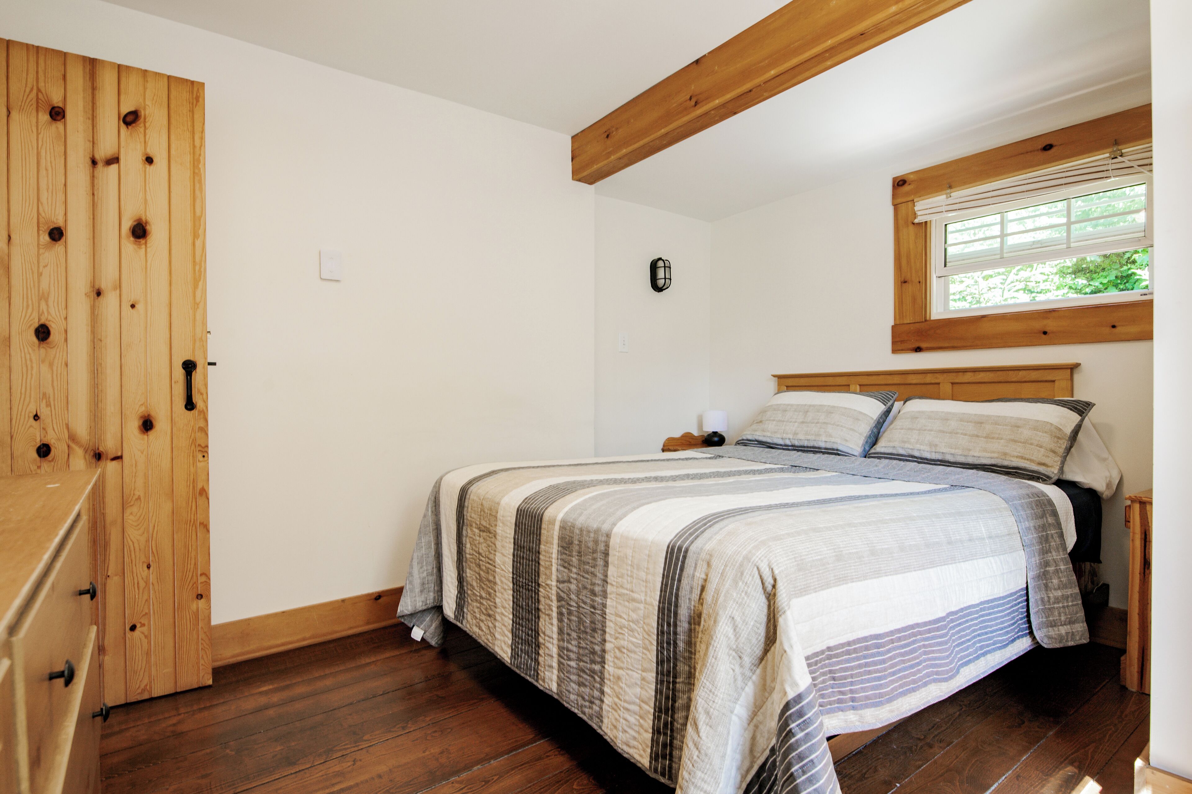 Un dormitorio con un armario de madera, una cama con colcha a rayas, una mesita de noche y una ventana con persianas.