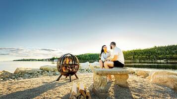 A couple sitting on a stone bench near a fire pit by a lake with a forested island in the background.