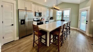 A spacious kitchen with a central island, white cabinets, stainless steel appliances, and a large window providing natural light. - Oyster & Pearl Beautiful New Single Level Condo (Corolla)