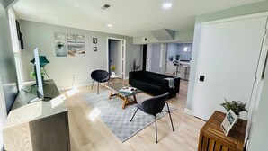 A modern living room with a black sofa, two black chairs, a coffee table, and a TV on a wooden stand. There is a kitchen area in the background with a bar stool and a plant.