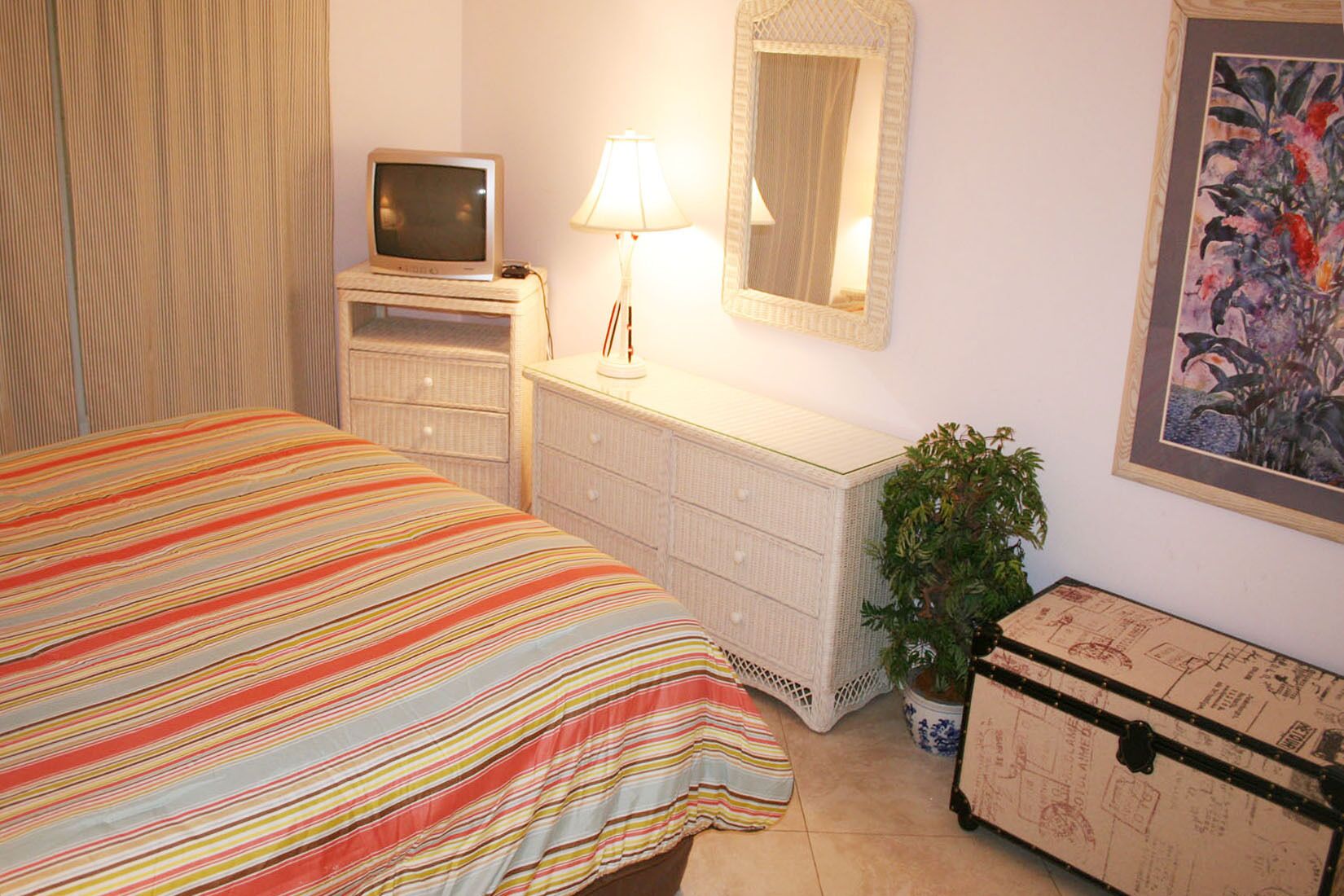 A bedroom with a bed, a dresser, a lamp, a television, a potted plant, and a suitcase.