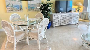 Un salon avec une table à manger en verre, des chaises en osier blanches, un téléviseur posé sur un meuble blanc et deux tableaux accrochés au mur.