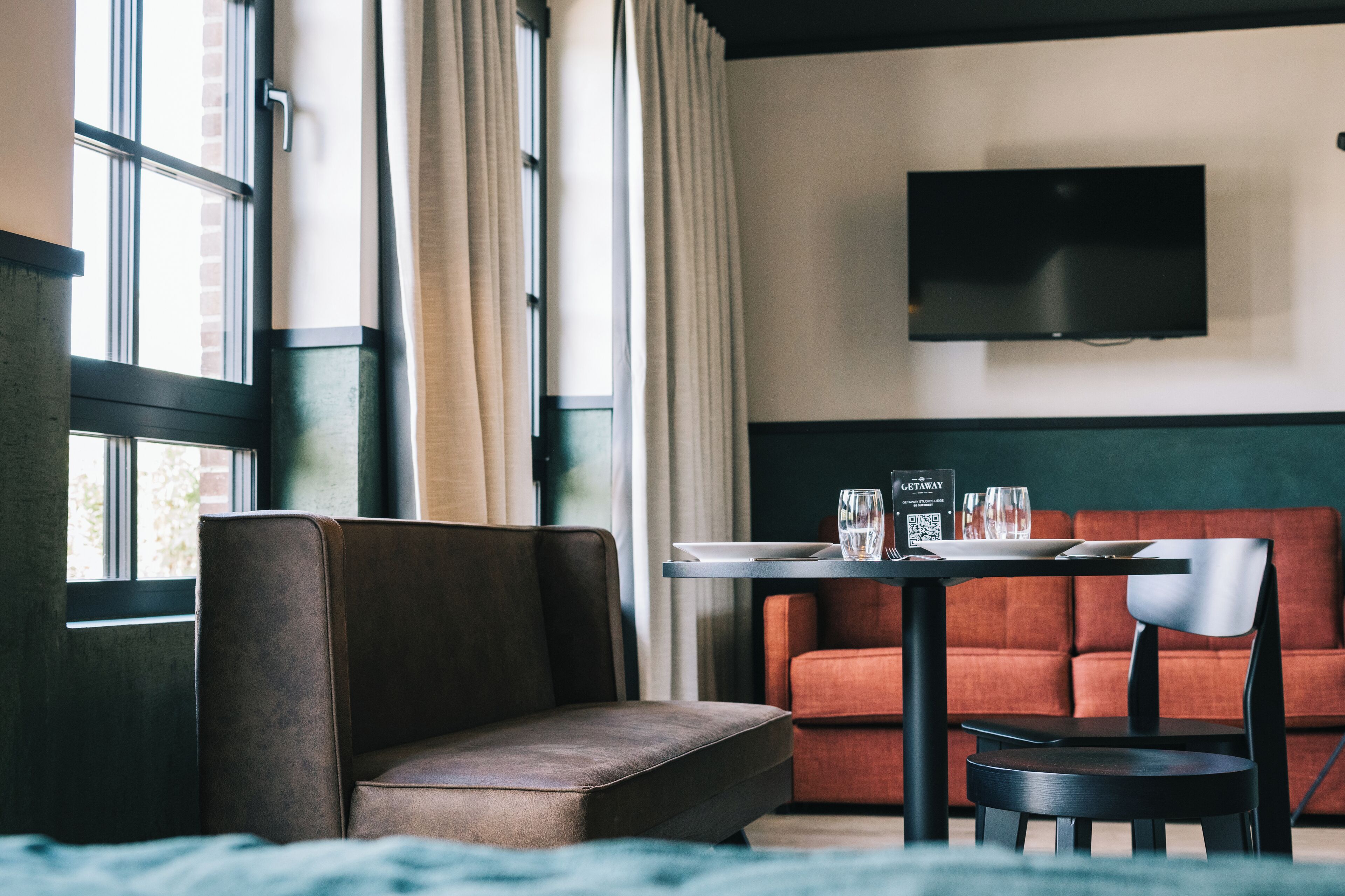 Una habitación de hotel moderna con un sofá, una mesita con un menú, un televisor de pantalla plana montado en la pared y ventanas grandes con cortinas.