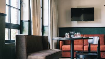 Una habitación de hotel moderna con un sofá, una mesita con un menú, un televisor de pantalla plana montado en la pared y ventanas grandes con cortinas.