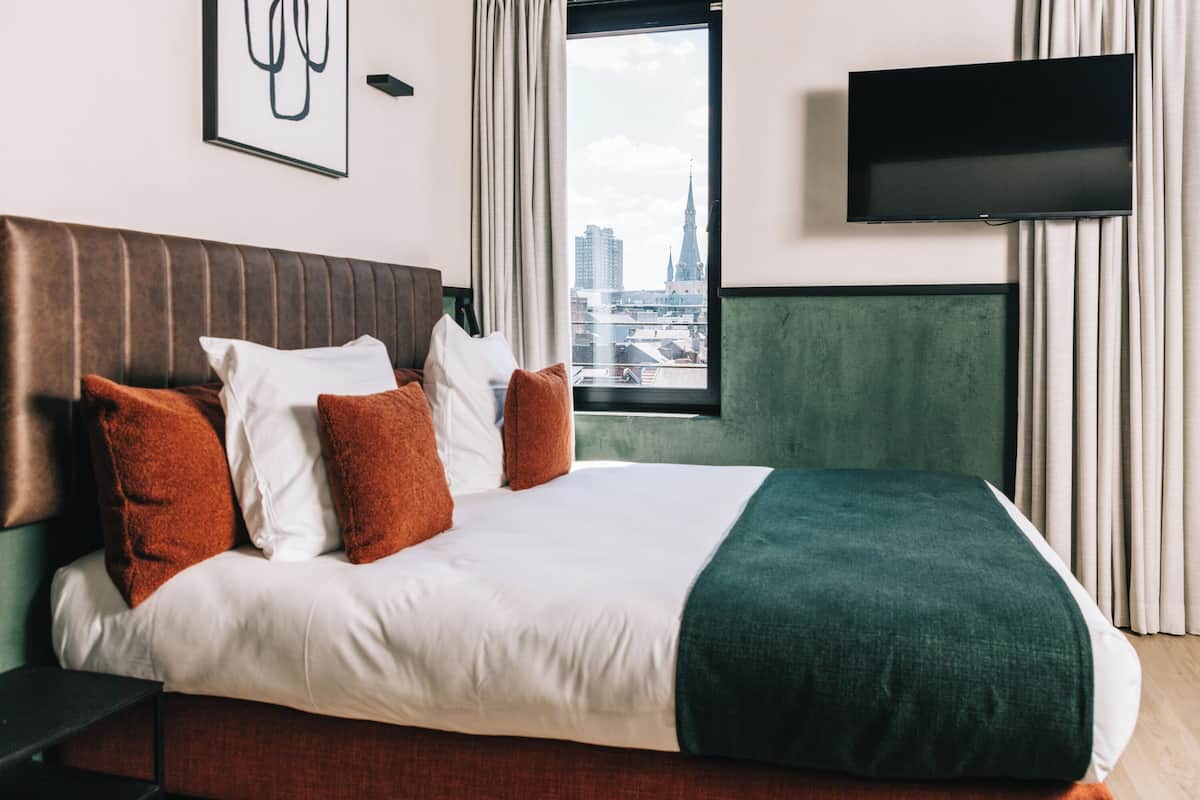 Ein Hotelzimmer mit einem großen Bett, einem Flachbildfernseher, Blick auf die Stadt und einem gerahmten Kunstwerk an der Wand.